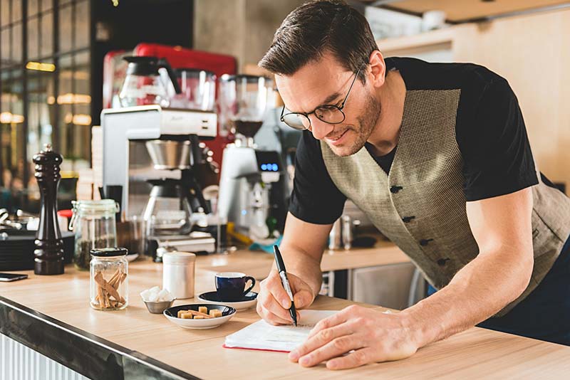 Male coffee shop owner stands over a counter and writes his small business strategy.