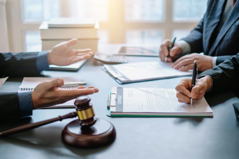 Two professionals in suits sign contracts at a desk next to a judge's gavel.